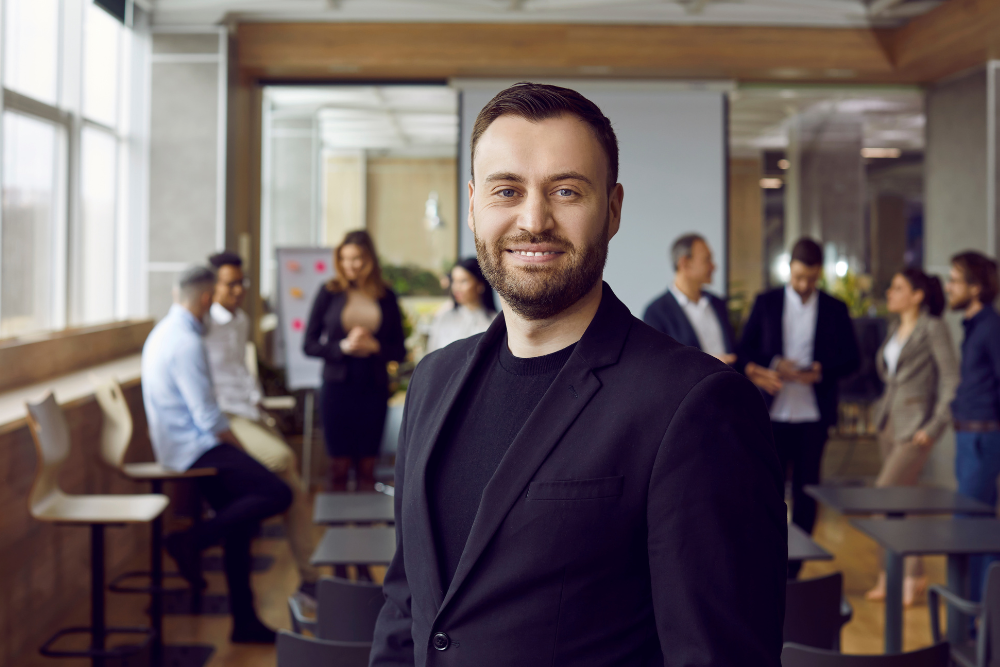 Selbstbewusster Geschäftsmann im Vordergrund lächelt in einem gut beleuchteten Büro mit Teammitgliedern, die im Hintergrund interagieren, was Team Performance Coaching und kollaborative Arbeitsumgebung darstellt.