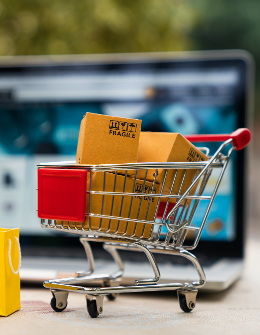 Ein Miniatur-Einkaufswagen gefüllt mit kleinen Paketen, auf denen "FRAGILE" steht, vor einem Laptop mit einer geöffneten Online-Shopping-Website. Der Einkaufswagen steht auf einer Oberfläche mit unscharfem Hintergrund, der grüne Pflanzen zeigt, was darauf hindeutet, dass das Bild in einem Innenraum mit Blick nach draußen aufgenommen wurde. Auf der linken Seite des Einkaufswagens ist eine kleine, unscharfe, gelbe Einkaufstüte zu erkennen.